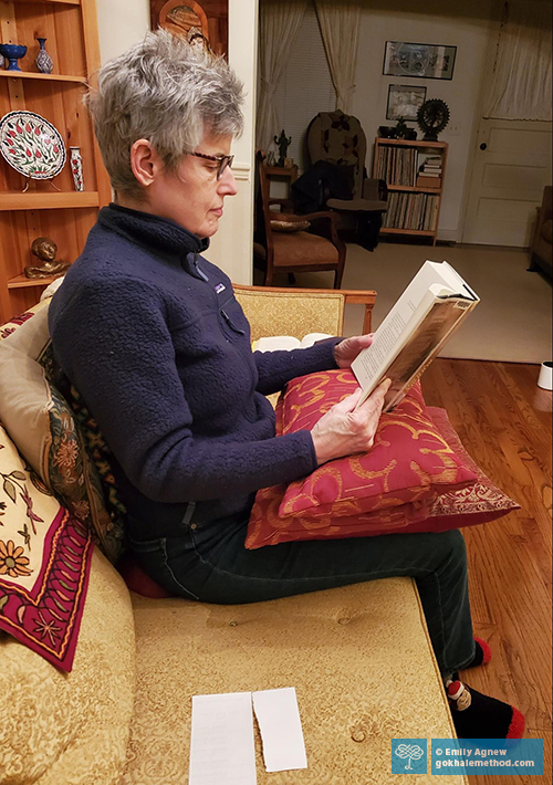 Photo of Emily Agnew reading, stretchsitting with supportive cushions on her couch.